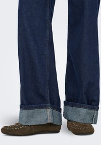 Jean en denim bleu foncé avec revers retroussés dévoilant un denim plus clair en dessous. Associé à des chaussures en daim marron avec des coutures décoratives.