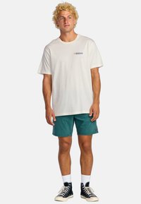 Camiseta blanca de algodón con un pequeño logo, combinada con pantalones cortos texturizados en tono verde azulado y zapatillas negras de caña alta con cordones blancos.