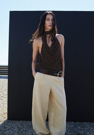 Femme aux longs cheveux foncés portant un haut noir sans manches à pois et un pantalon beige large avec une ceinture noire, debout devant un fond sombre.