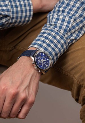 Montre bleue avec accents argentés ; cadran rond affichant trois sous-cadrans, fonction date et chiffres. Brigitte en cuir bleu au motif crocodile.