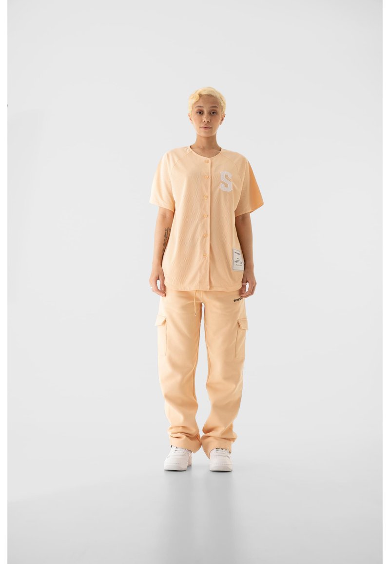 Őszibarack színű, hálós anyagú gombos felső fehér "S" betűvel, hozzáillő cargo nadrággal, zsebekkel és laza szabással, fehér tornacipővel párosítva.
