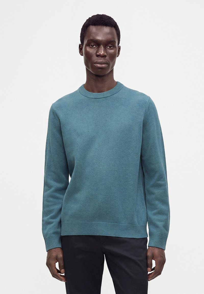 Modèle masculin portant un pull col rond bleu sarcelle et un pantalon noir, debout sur un fond blanc uni.