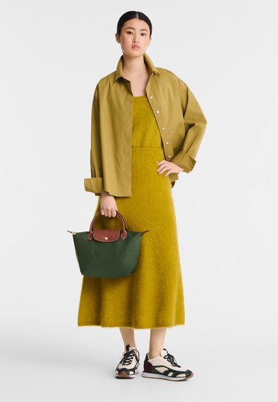 Olivgrünes, übergroßes Hemd, senfgelbes Kleid, dunkelgrüne Tasche mit braunen Akzenten und neutrale Sneaker mit schwarzen Schnürsenkeln und strukturierten Details.