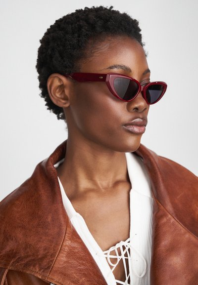 Maroon cat-eye sunglasses with dark lenses and glossy finish. Accompanied by a brown leather jacket and white top with lacing detail.
