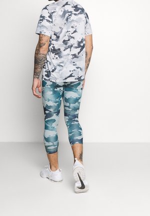 Mężczyzna z tatuażami na ramionach i nogach, ubrany w białą szaro-moro koszulkę, niebieskie moro legginsy do kolan oraz białe buty sportowe, stoi tyłem.