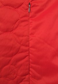 Röd quiltad tyg med ett mönster av cirkulära stygn, med en slät dragkedja på ena sidan. Matt finish och lätt textur.