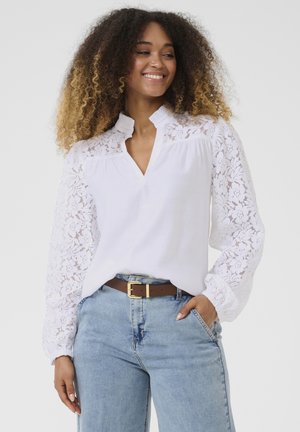 White blouse with lace long sleeves, a mandarin collar, and a relaxed fit. Paired with blue jeans and a brown belt with a gold buckle.