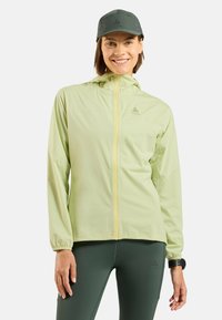 Light green windbreaker jacket with a hood and yellow zipper, paired with dark green leggings and a gray cap. Features elastic cuffs.