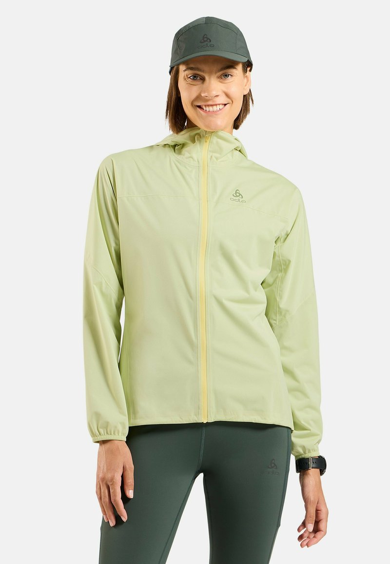 Light green windbreaker jacket with a hood and yellow zipper, paired with dark green leggings and a gray cap. Features elastic cuffs.