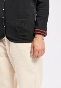 Schwarze Knopf-Cardigan mit abgerundetem Saum, der eine geraffte Tasche und rot gestreifte Ärmelbündchen aufweist. Mit cremefarbenen, weit geschnittenen Hosen kombiniert.