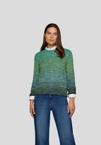 Eine Frau mit langen braunen Haaren trägt einen grün-blauen Farbverlauf-Strickpullover über einem weißen Hemd und blauen Jeans und steht vor einem schlichten grauen Hintergrund.