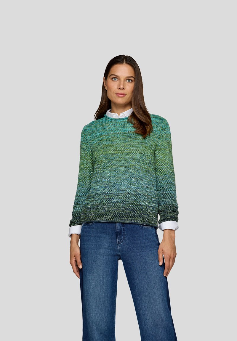 Eine Frau mit langen braunen Haaren trägt einen grün-blauen Farbverlauf-Strickpullover über einem weißen Hemd und blauen Jeans und steht vor einem schlichten grauen Hintergrund.