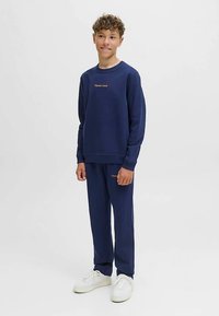 Tmavomodrá mikina a nohavice z mäkkého materiálu; majú okrúhly výstrih, dlhé rukávy a zlatú vyšívanú značku na hrudi a stehne.