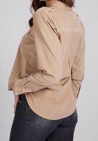 Femme portant une chemise beige à manches longues avec des poignets boutonnés et un jean noir, légèrement tournée pour montrer les détails du dos et du côté du vêtement.