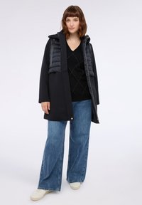 Schwarzer Mantel mit Kapuze und puffy grauer Westen-Overlay, kombiniert mit einem schwarzen V-Ausschnitt-Pullover und weiten blauen Jeans. Weiße Sneakers runden den Look ab.