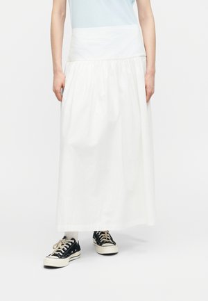 Oseba, ki nosi svetlo modro zgornji del obleke, dolgo belo krilo, bele nogavice in črno nizko športno obutev, stoji na enakomerni podlagi.