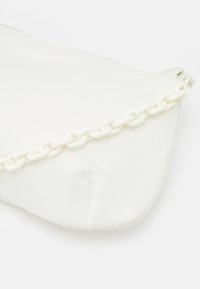 Sac blanc à texture lisse, avec un accent de chaîne blanche brillante le long du bord supérieur et une fermeture éclair. Forme incurvée et asymétrique.