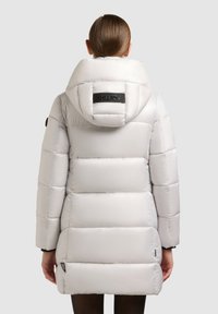 Pufjakke i lysegrå materiale, der har en stor hætte og blokkviltet mønster. Sort logoaccents på ryggen og lynlåslommer i siderne.