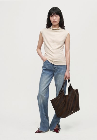 Un mannequin se tient devant un fond uni, vêtu d’un haut crème drapé sans manches, d’un jean à jambes larges et de ballerines bordeaux, et tient un grand cabas marron.