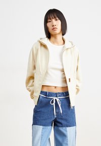 Cream zip-up hoodie with side pockets; paired with denim shorts featuring varying shades of blue and a white drawstring. Smooth texture.