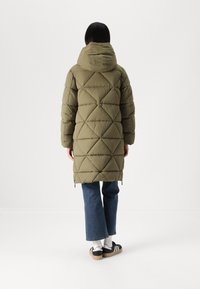 Olivgrüner Puffer-Mantel mit Kapuze, der mit einer Rautensteppung und einer etwas längeren Passform versehen ist, kombiniert mit weit geschnittenen Jeans und sportlichen Schuhen.