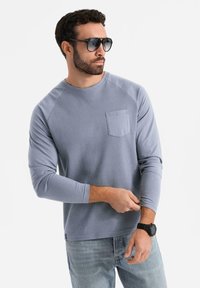 Heller blauer Langarmshirt mit strukturiertem Stoff, Rundhalsausschnitt und Brusttasche. Kombiniert mit dunklen Sonnenbrillen und blauen Jeans.