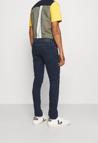 Jeans de mezclilla oscura ajustados, combinados con una camisa de manga corta multicolor, que presenta paneles de tela en contraste y zapatillas blancas con acentos.