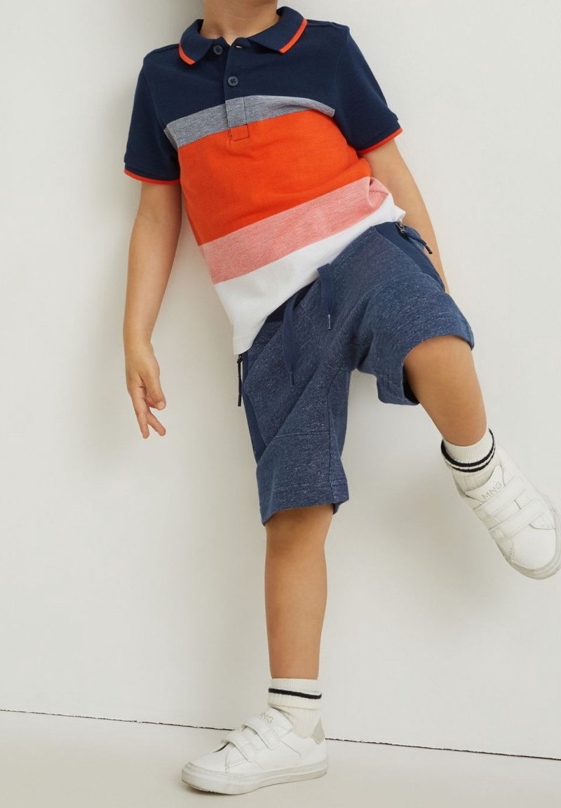 Enfant portant un polo rayé bleu marine, orange, rose et blanc avec un short bleu marine, levant une jambe en restant debout contre un mur blanc.