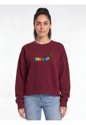 Femme portant un sweat-shirt bordeaux avec un teckel rayé coloré tenant une fleur jaune devant, associé à un jean bleu clair.