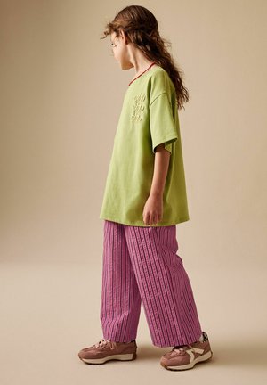 Child with long hair wearing a green "ciao" shirt, pink striped wide-leg pants, and pink sneakers standing sideways on a beige background.