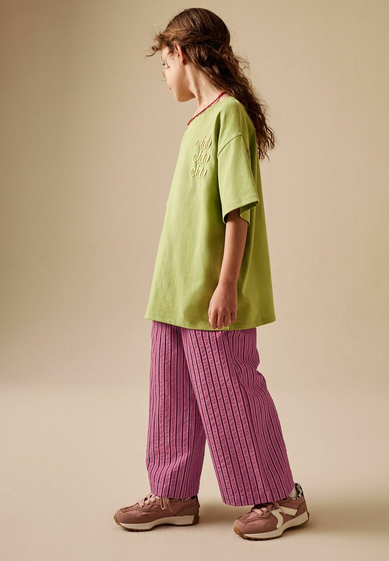 Hosszú hajú gyermek zöld "ciao" feliratú pólóban, rózsaszín csíkos bő szárú nadrágban és rózsaszín sportcipőben, oldalirányban áll egy bézs háttér előtt.