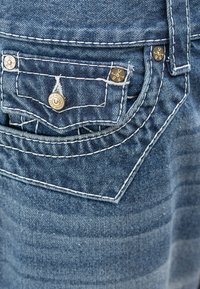 Κοντινή λήψη τσέπης από μπλε τζιν με λευκό ράψιμο και μεταλλικά καρφιά σε μικρό καπάκι τσέπης.