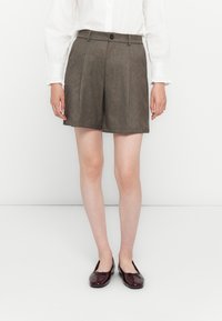 Witte blouse met lange mouwen in de bruine op maat gemaakte shorts gestoken, gedragen met donkerburgundy ballerina's op een witte achtergrond.