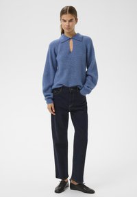 Modra pletena jopica s ovratnikom in detajlom v obliki ključavnice, kombinirana s temno modrimi naravnost krojenimi kavbojkami in črnimi čipkastimi čevlji.