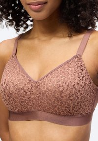 Γυναίκα που φοράει μπραλέτα από δαντέλα σε μωβ απόχρωση με ρυθμιζόμενες τιράντες, υφασμένο λουλουδάτο σχέδιο και φαρδιά ελαστική ζώνη κάτω από το στήθος.