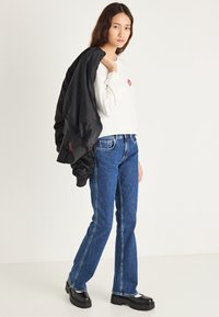 Cazadora de aviador negra colgada sobre un hombro, sudadera blanca con un logo rojo, vaqueros acampanados azules y botas negras gruesas. Texturas suaves.