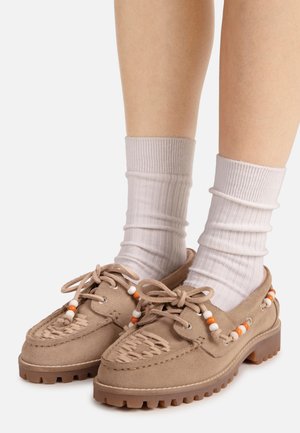 Jambes portant des mocassins en daim beige clair avec des semelles épaisses et des chaussettes blanches, ornés de perles orange et blanches sur les lacets et les côtés.