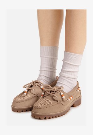 Jambes portant des mocassins en daim beige clair avec des semelles épaisses et des chaussettes blanches, ornés de perles orange et blanches sur les lacets et les côtés.
