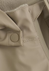 Beige, waterdichte stof met gestikte naden. Voorzien van een ronde knop met reliëflogo. Zachte, gestructureerde voering zichtbaar aan de rand.