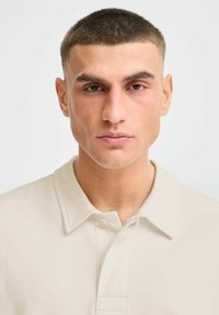 Polo beige au design classique, avec un col boutoné et une texture lisse, porté par une personne ayant les cheveux très courts.