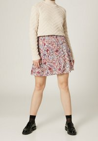 Suéter de punto beige con un cuello alto y un patrón de zigzag, combinado con una falda mini de estampado floral en tonos rosas y azules. Mocasines negros y calcetines.