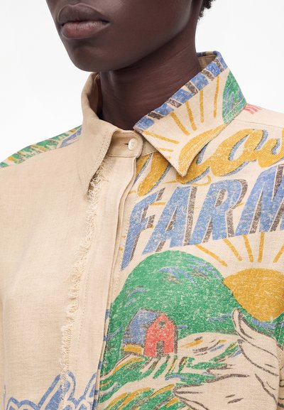 Άτομο που φοράει μπεζ πουκάμισο με κουμπιά, με πολύχρωμα εκτυπωμένα γραφικά θεματολογίας αγροκτήματος και φθαρμένο ραφικό σχέδιο κοντά στον εμπρός καβάλο.