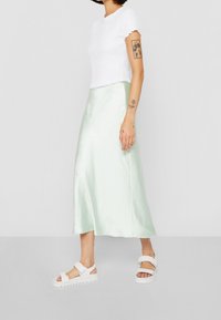 Kvinna i en vit figurnära topp med kort ärm, ljusgrön satinkjol i midi-längd, vita sandaler, och tatueringar på arm och handled, stående i profil.