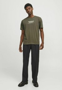 Olivno zelena majica s kratkimi rokavi in okroglo izrezom, ki vsebuje bel tisk. V kombinaciji z črnimi nabranimi hlačami in črnimi čevlji.