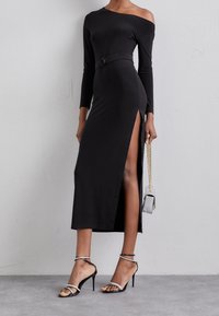 Černé šaty s dlouhým rukávem a designem bez ramínek, s bočními rozparky. Obsahují přiléhavý pás a jsou spárované s černými sandály na tenkých podpatcích.