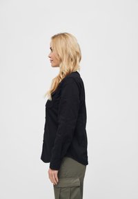 Camicia nera a maniche lunghe con colletto, dotata di tasche con bottoni; abbinata a pantaloni cargo verde oliva; vista di profilo.