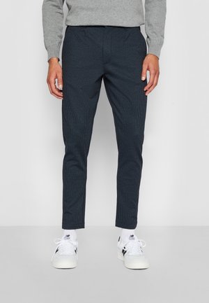 Mand iført mørke slim-fit bukser, lys grå sweater, hvide sokker og hvide sneakers med sorte detaljer, stående mod en ensfarvet baggrund.