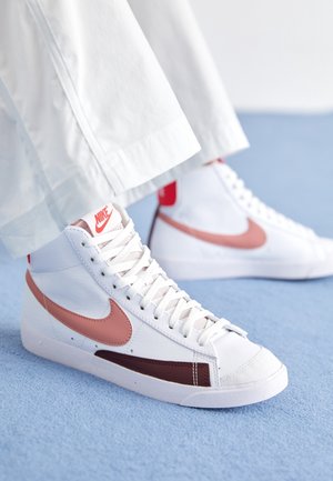 Valkoiset korkeavartiset Nike-tennarit, joissa on vaaleanpunainen swoosh ja ruskea yksityiskohta, käytettyinä valkoisten housujen kanssa vaaleansinisellä matolla.