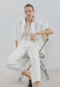 Mlada ženska s svetlimi lasmi, zvezanimi nazaj, sedi na kovinskem stolu, oblečena v belo srajco, bele hlače, brezrokavnik, črne gležnjarje in uhani s privezakom.