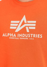 T-shirt in cotone arancione con logo bianco caratterizzato da un design astratto e il testo "ALPHA INDUSTRIES KNOXVILLE, TENNESSEE, U.S.A."